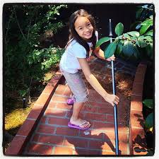 A little girl smiling at the camera while she is sweeping a sidewalk