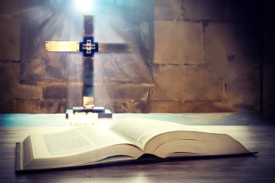 Bible on table with cross and light beaming through a window behind shining on the cross