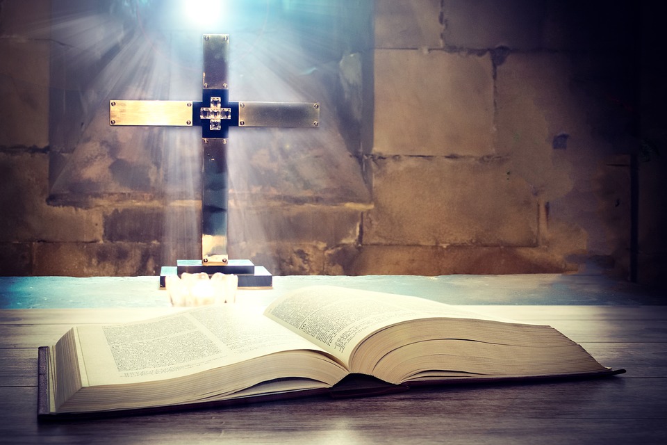 open Bible with sunlight streaming in and reflecting off a golden cross behind the Bible