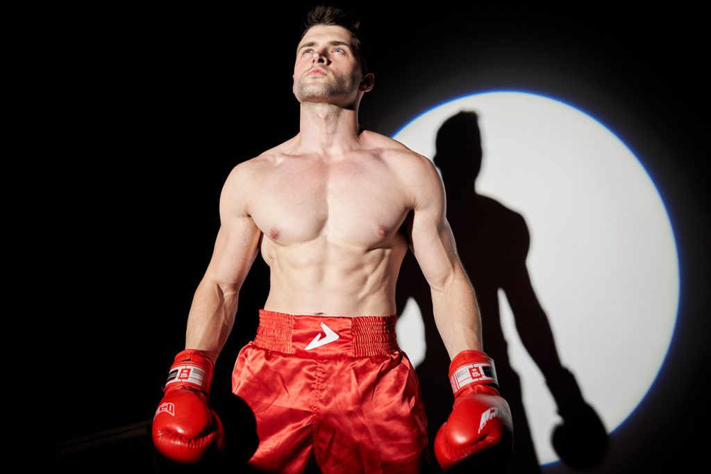 boxer in red shorts with red globes standing in the spotlight looking determined