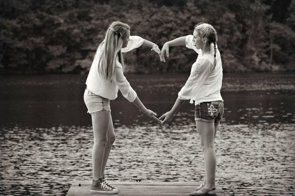 two girls using arms to make heart standing on the dock of a lake