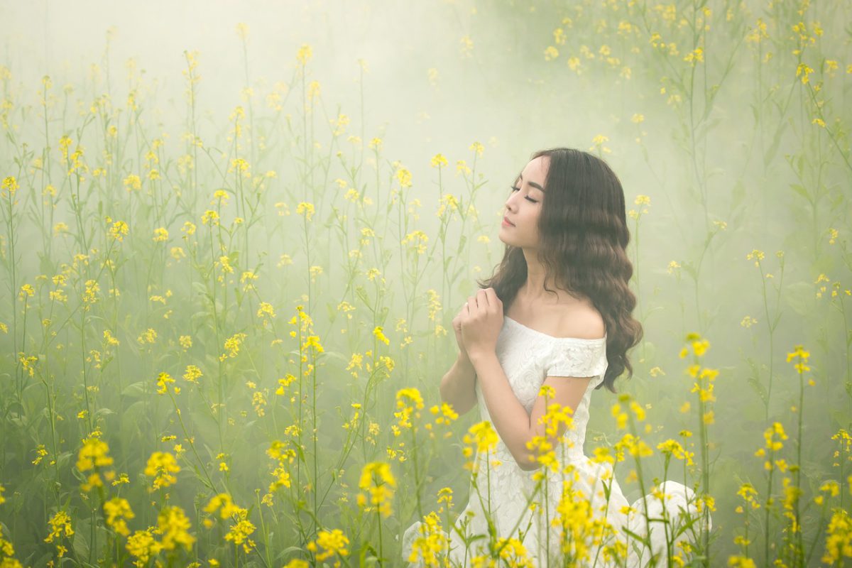 girl in white dress amidst yellow flowers in a field, with praying hands and mist around her