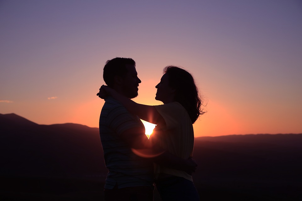 a man and woman in light embrace, facing each other, with a sunset behind them