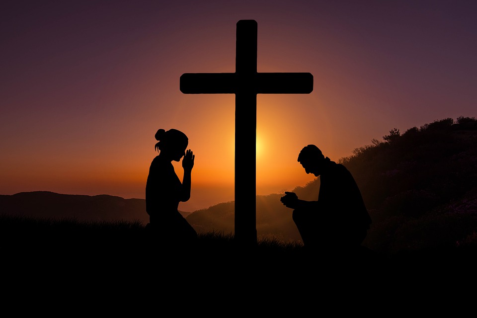 silhouette of a man and a woman kneeling at the foot of a cross with a sunset in the background