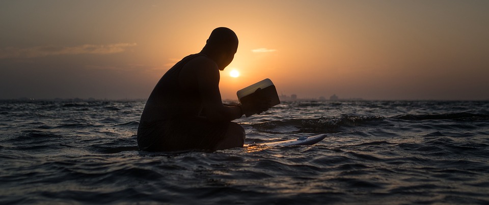 man sitting in water, reading with the sunset behind him